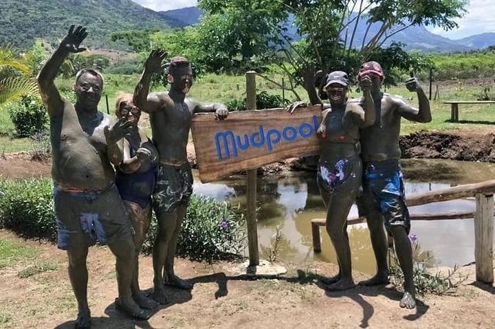 Visitors covered in mud waving beside the Sabeto mud pool sign